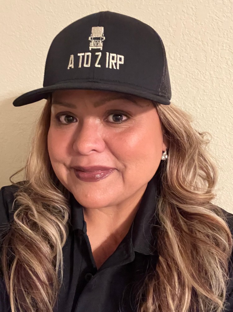 A woman with long, wavy hair wearing a black cap featuring the text 'A TO Z IRP', and a black shirt. She is smiling and poses against a light-colored wall.